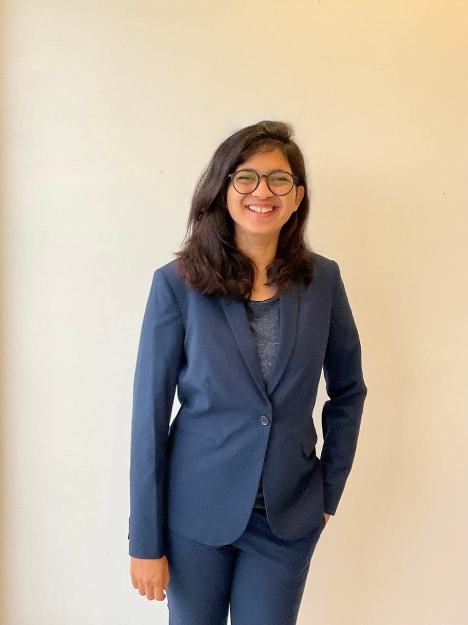 Saloni Girhepuje smiling at camera - navy blazer over a heathered grey top, round glasses, against an off-white wall