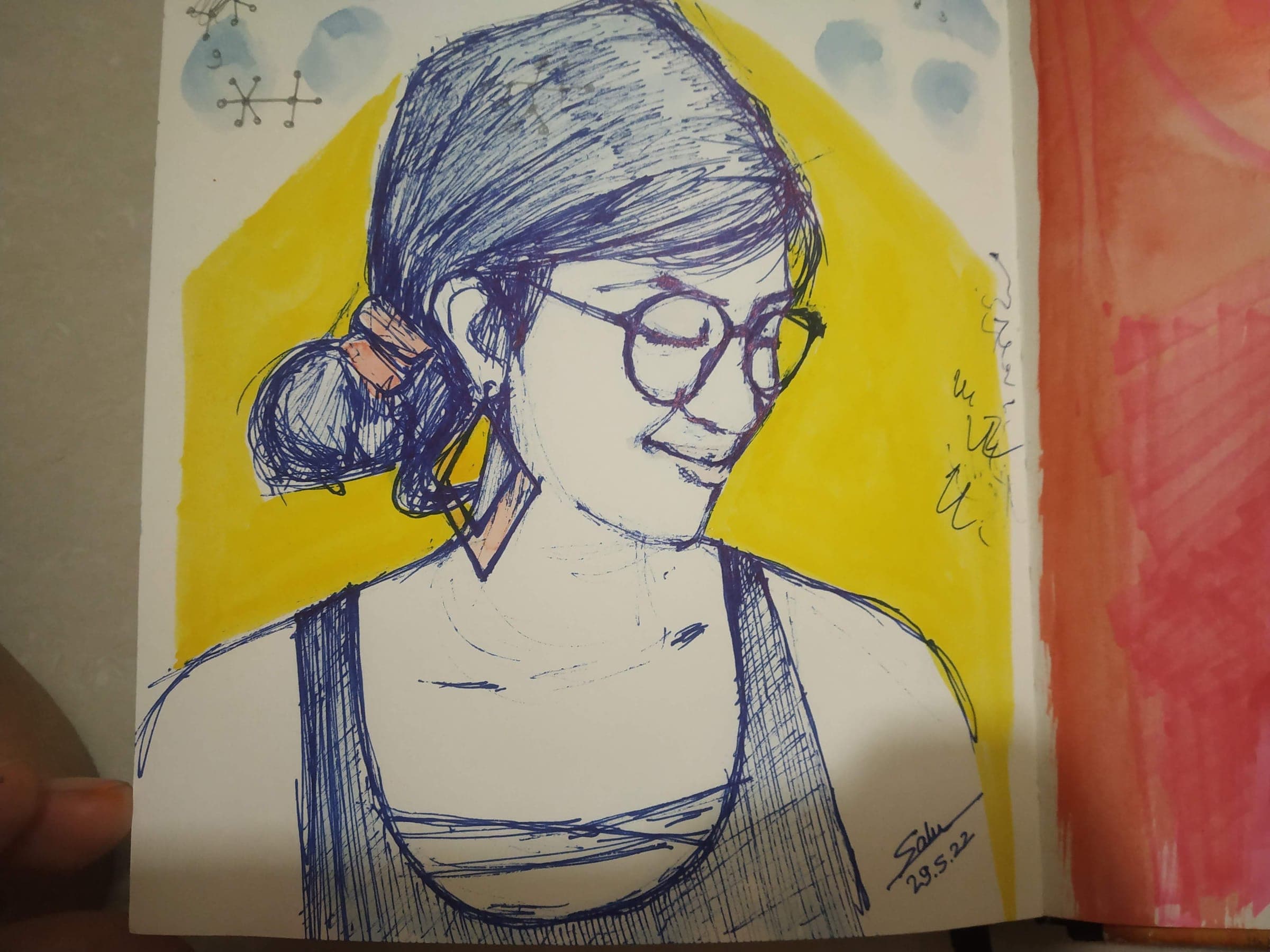 Self-portrait in blue ballpoint pen over a yellow watercolor wash: a smiling woman with glasses and a messy bun, eyes closed, signed Salu 29.5.22 in the corner of the sketchbook page