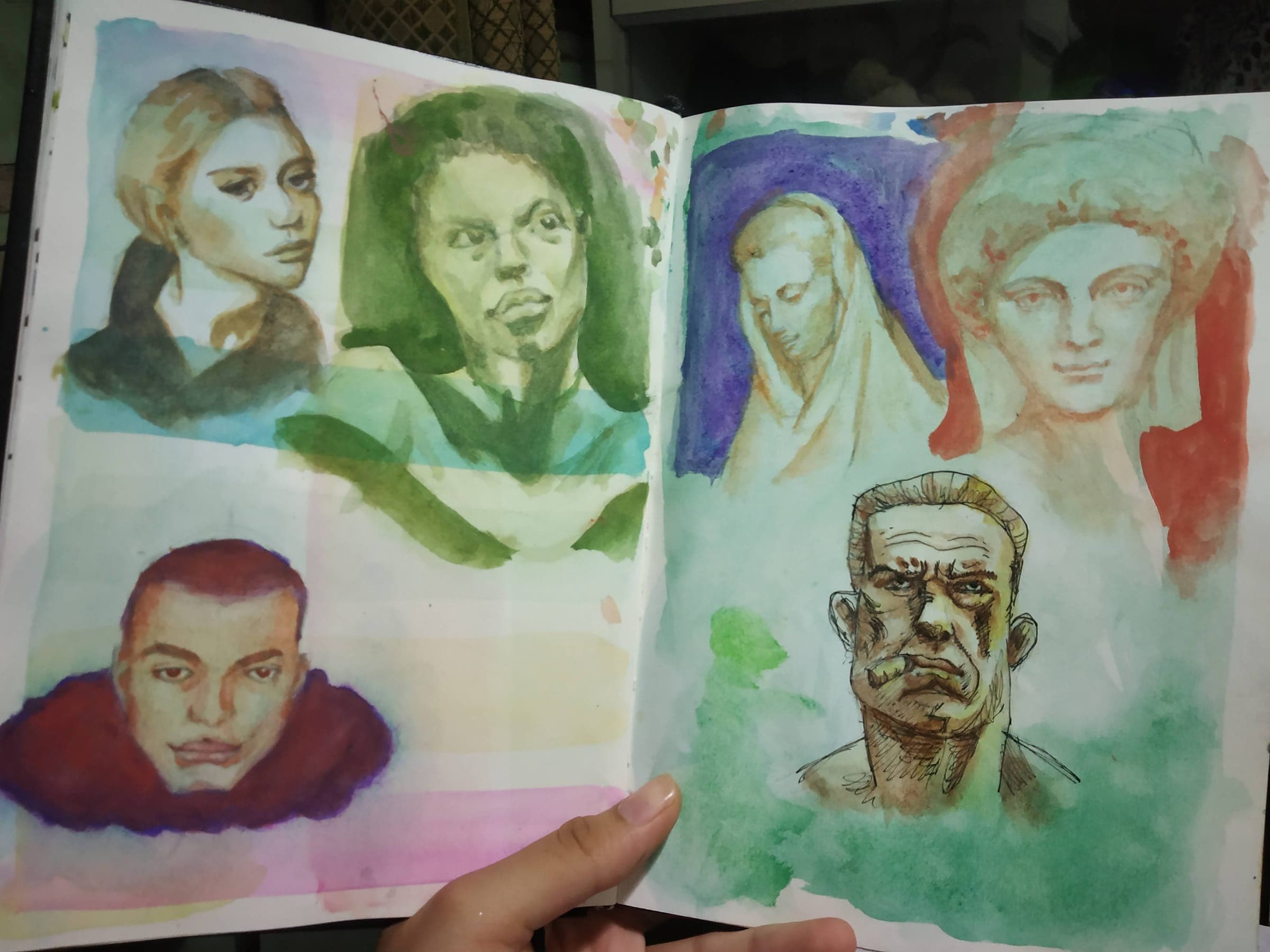 An open sketchbook spread holding six loose watercolor portrait studies: a blonde woman, a green-toned frowning face, a red-hooded head, two classical statue heads in veils, and an ink-and-wash old man